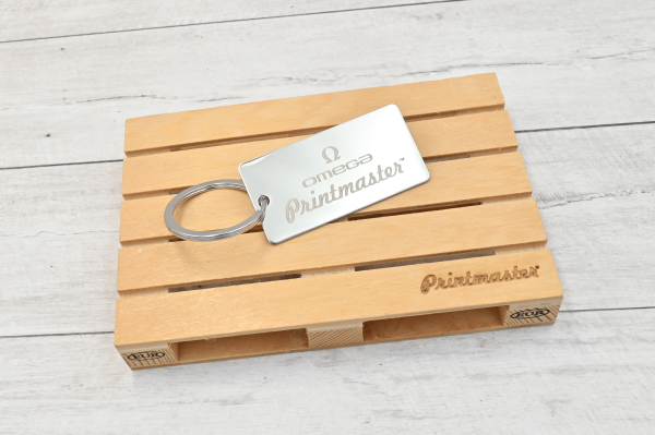 Llavero tipo tag rectangular en acero inoxidable grabado con láser, con logo de Omega Printmaster sobre base de madera.