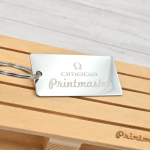 Llavero tipo tag rectangular en acero inoxidable grabado con láser, con logo de Omega Printmaster sobre base de madera.