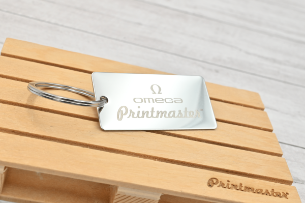 Llavero tipo tag rectangular en acero inoxidable grabado con láser, con logo de Omega Printmaster sobre base de madera.