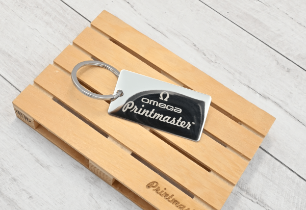 Llavero tipo tag rectangular en acero inoxidable grabado con láser, con logo de Omega Printmaster sobre base de madera.