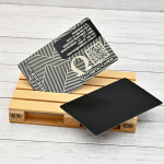 Tarjeta metálica grabada con láser Omega Láser, con diseño geométrico, logo Omega y código QR, sobre fondo de madera clara.