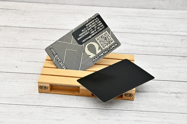 Tarjeta metálica grabada con láser Omega Láser, con diseño geométrico, logo Omega y código QR, sobre fondo de madera clara.