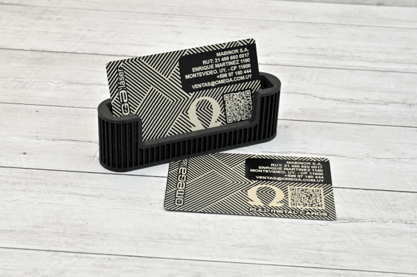 Tarjeta metálica grabada con láser Omega Láser, con diseño geométrico, logo Omega y código QR, sobre fondo de madera clara.
