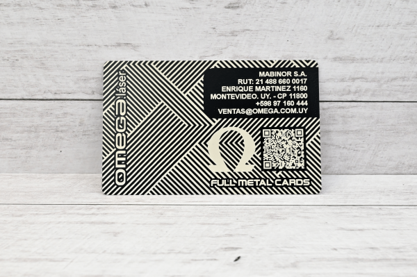 Tarjeta metálica grabada con láser Omega Láser, con diseño geométrico, logo Omega y código QR, sobre fondo de madera clara.