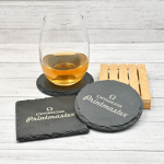 Posavasos de piedra natural grabados con láser con el logo Omega Printmaster, junto a un vaso con bebida sobre fondo de madera clara.