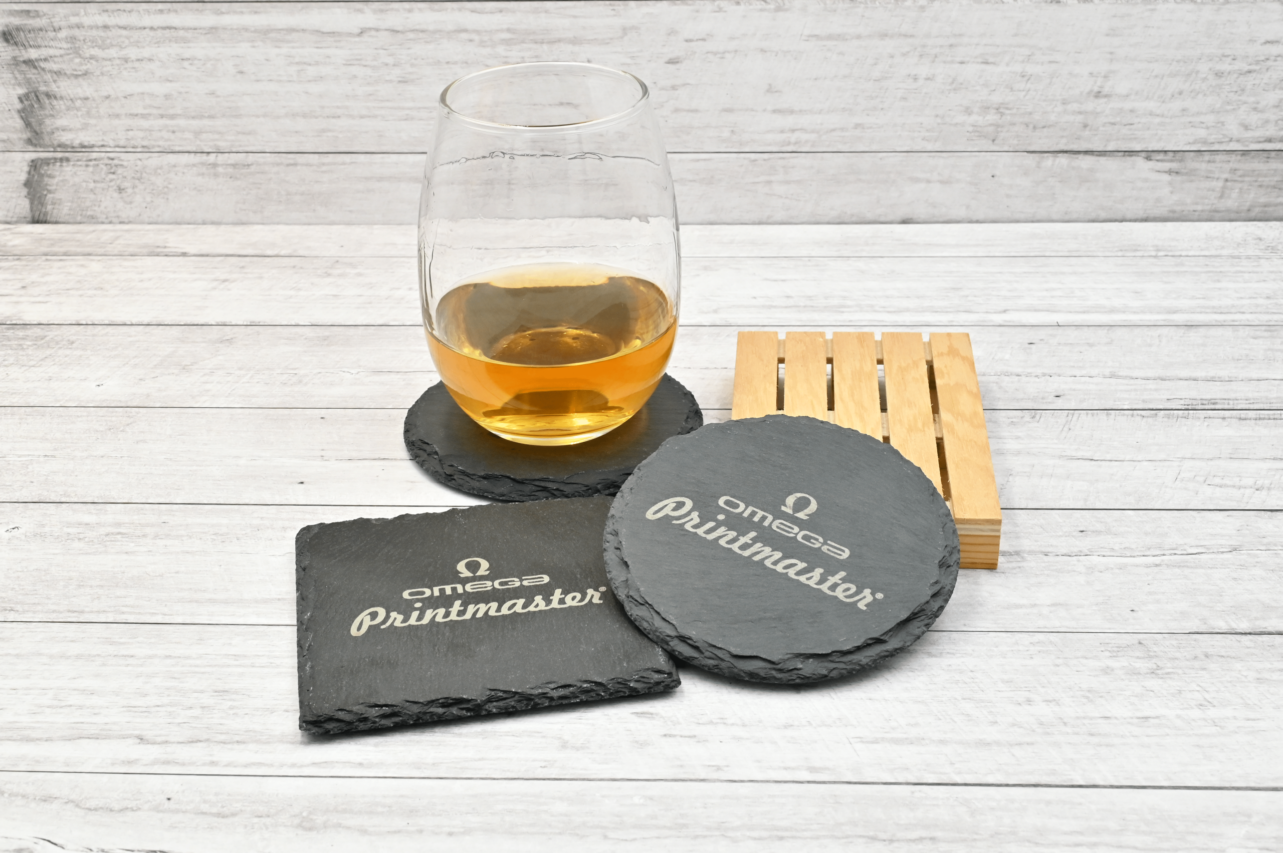 Posavasos de piedra natural grabados con láser con el logo Omega Printmaster, junto a un vaso con bebida sobre fondo de madera clara.