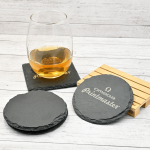 Posavasos de piedra natural grabados con láser con el logo Omega Printmaster, junto a un vaso con bebida sobre fondo de madera clara.