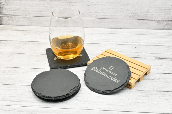 Posavasos de piedra natural grabados con láser con el logo Omega Printmaster, junto a un vaso con bebida sobre fondo de madera clara.