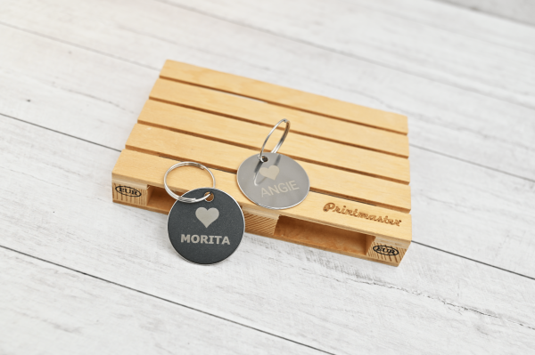 Dos tags circulares de acero inoxidable grabados con láser con los nombres “Morita” y “Angie”, apoyados sobre mini pallet de madera grabado con el logo Printmaster.