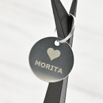 Tag redondo de acero inoxidable grabado con láser con el nombre “Morita” y un ícono de corazón, colgado sobre cinta negra.