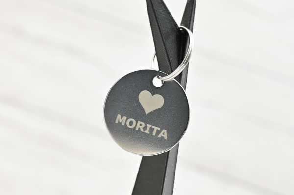 Tag redondo de acero inoxidable grabado con láser con el nombre “Morita” y un ícono de corazón, colgado sobre cinta negra.