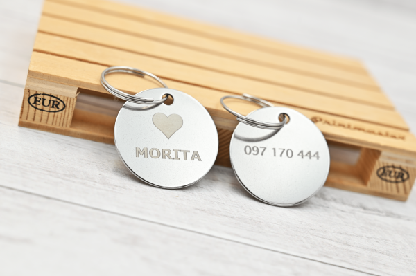 Tags circulares de acero inoxidable grabados con láser, con nombre y número personalizados, sobre mini pallet de madera con logo Omega Printmaster.