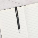 Lapicera Prestige Negra con logo Omega grabado a láser sobre cuaderno de hojas rayadas, fotografía de presentación.