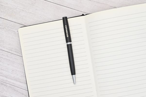 Lapicera Prestige Negra con logo Omega grabado a láser sobre cuaderno de hojas rayadas, fotografía de presentación.