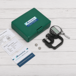 Kit completo del medidor digital de espesor con estuche rígido, baterías LR44, certificado de calibración y documentación sobre fondo de madera blanca.