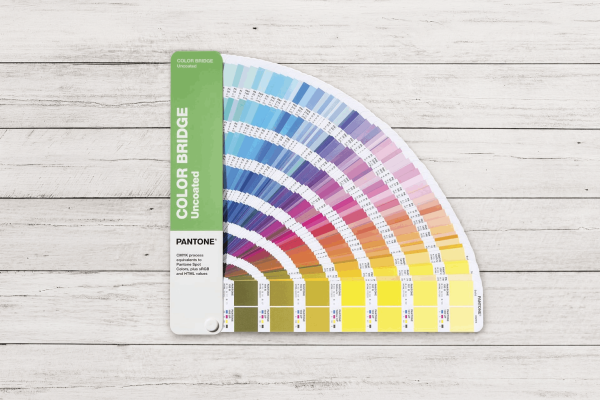 Guía Pantone Color Bridge Uncoated abierta en abanico sobre superficie de madera blanca, mostrando referencias Pantone y equivalencias CMYK para impresión profesional.